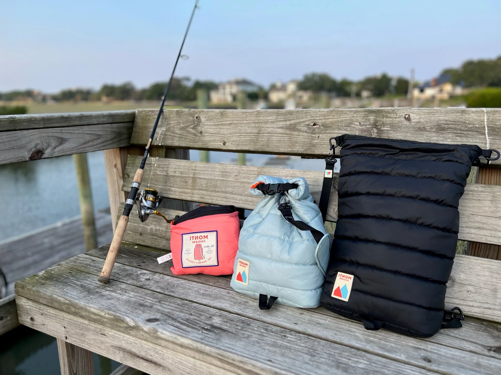 Fishing rod and three colorful bags on a wooden bench by a lake.