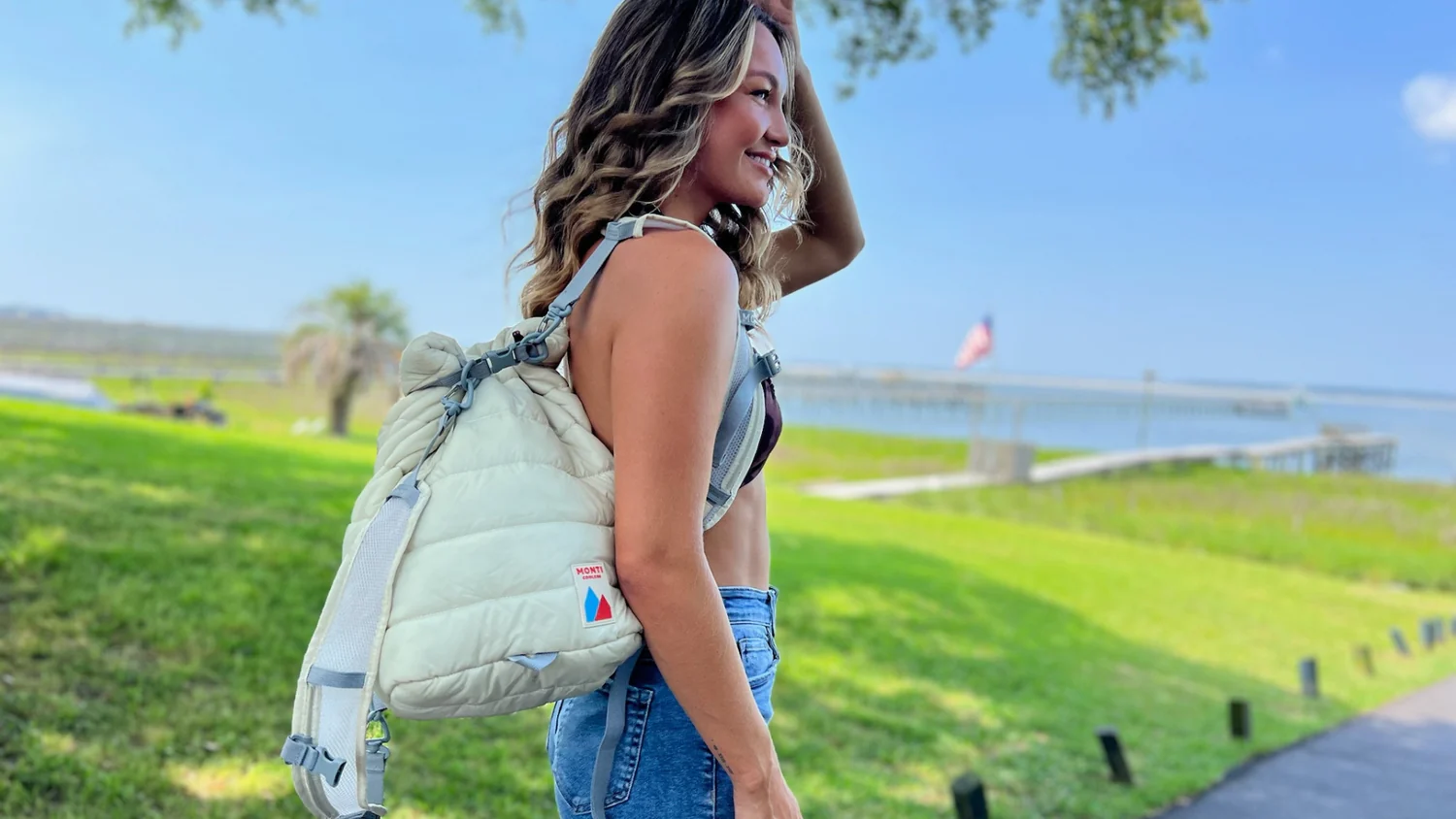 Person with puffy white backpack stands by grassy waterfront, smiling under a tree.