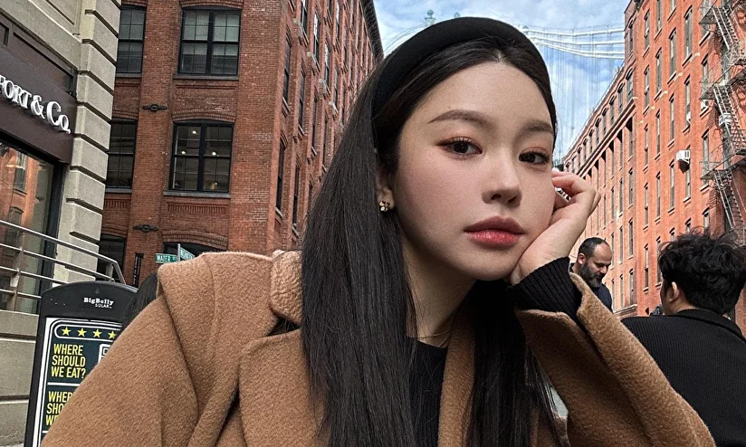 A woman in a brown coat and black headband poses on a city street with brick buildings.