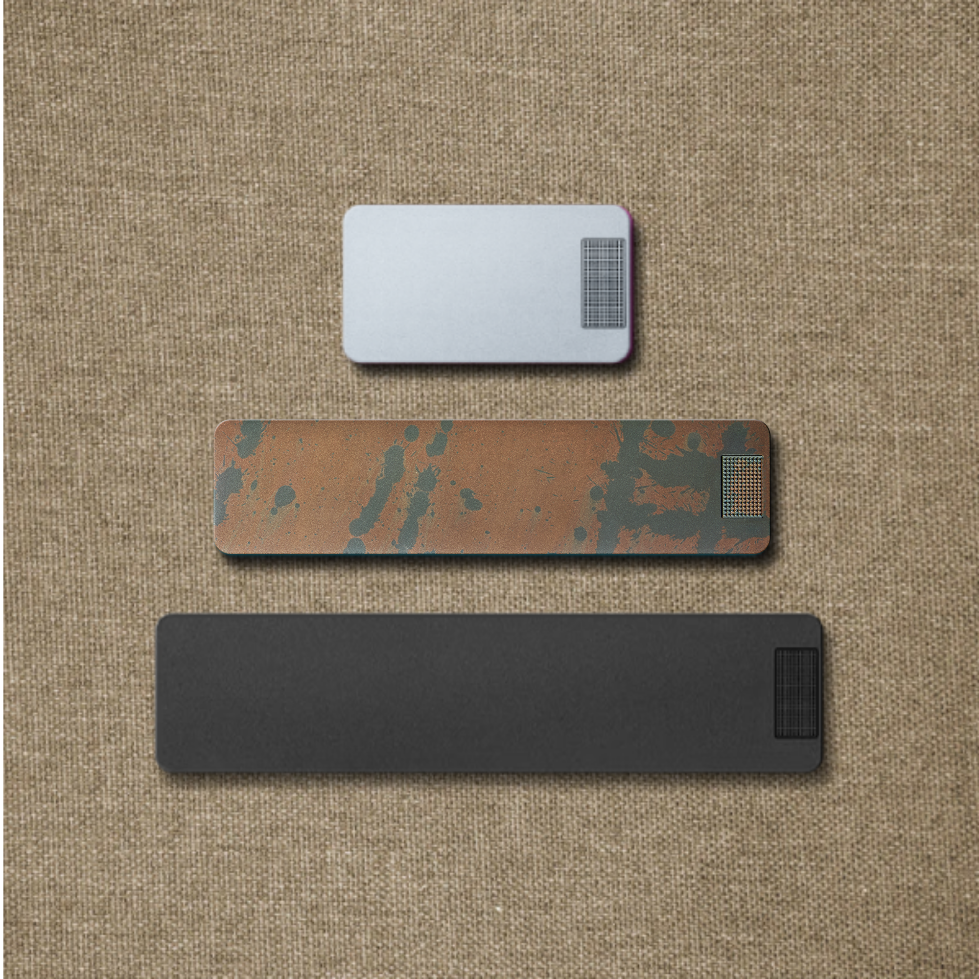 Three rectangular objects in silver, rust, and black finishes are stacked vertically on a burlap background.