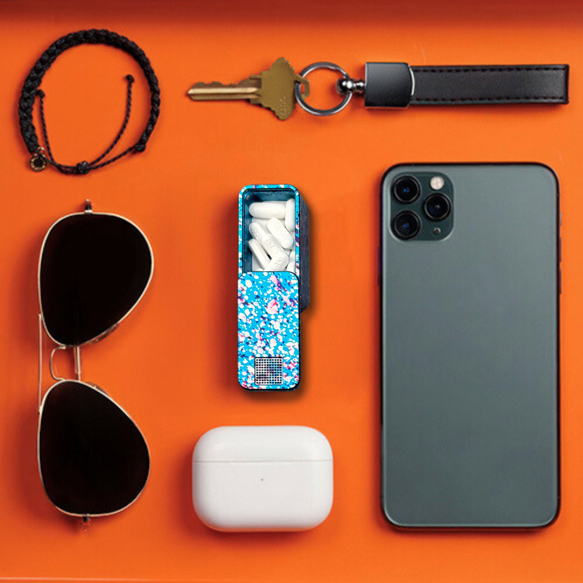 A flat lay of a smartphone, keys, sunglasses, pill case, and other personal items on an orange background.