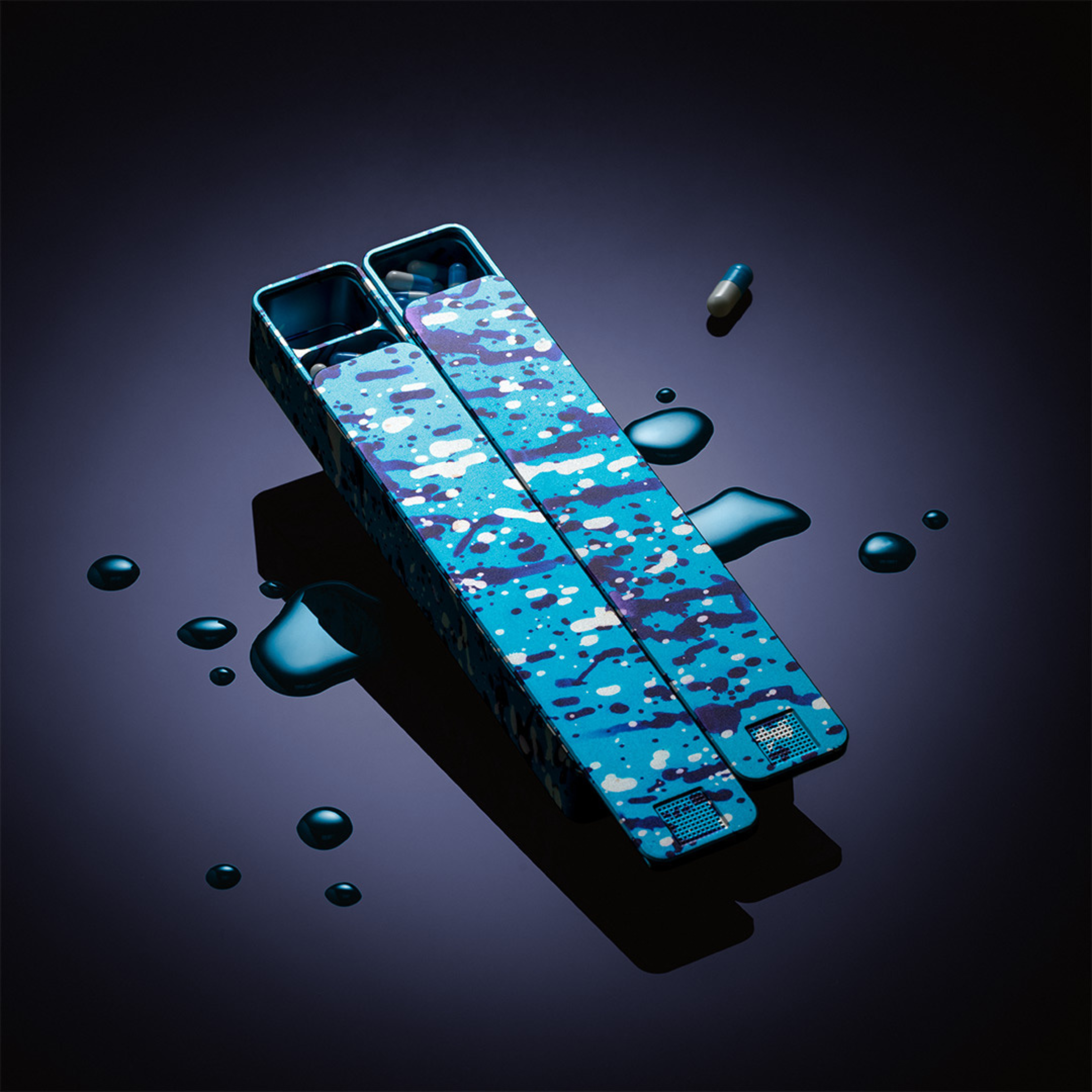 Two open, blue-patterned pillboxes with pills inside, surrounded by drops of liquid on a dark surface.