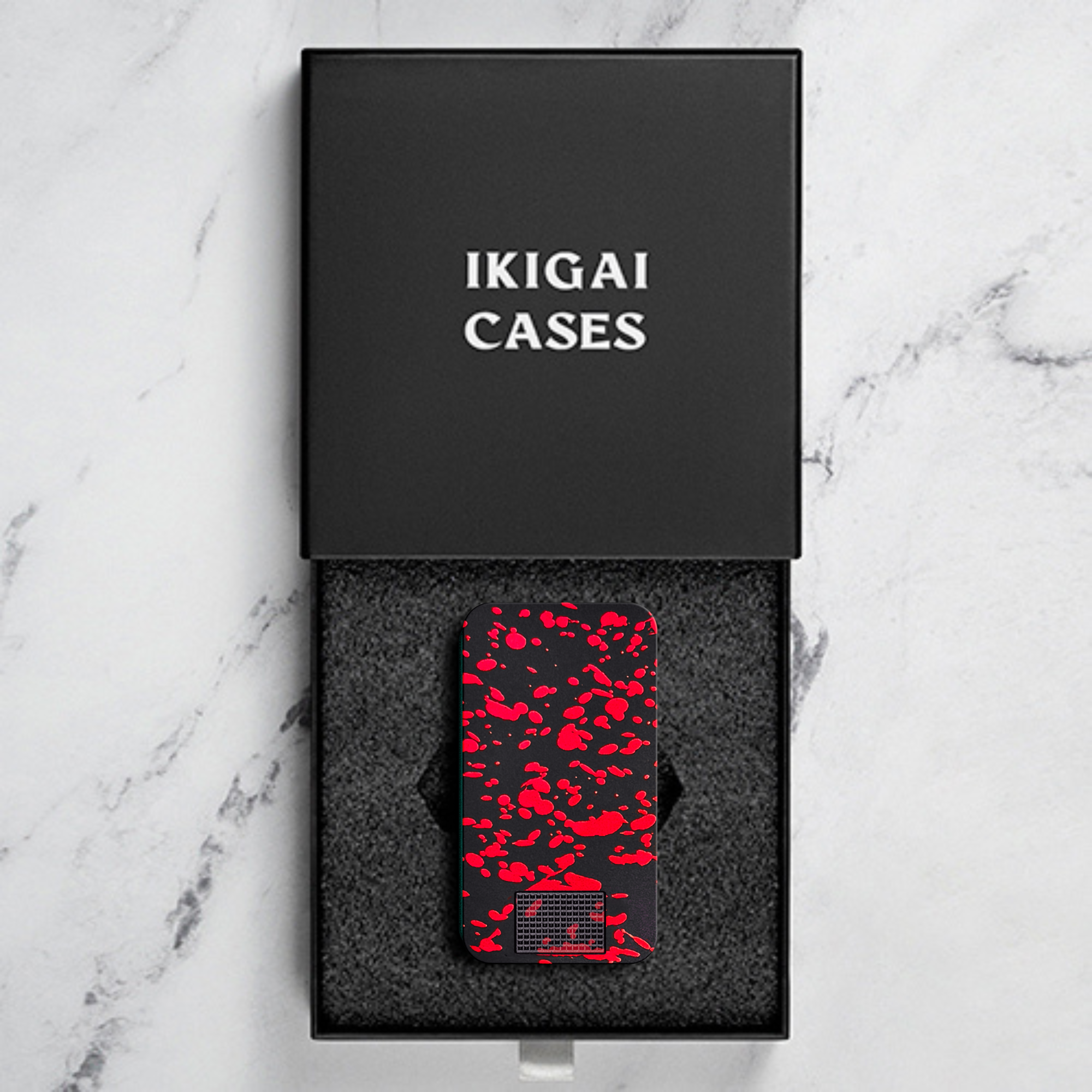 A black device with a red splatter pattern sits inside its open 'IKIGAI CASES' branded box.