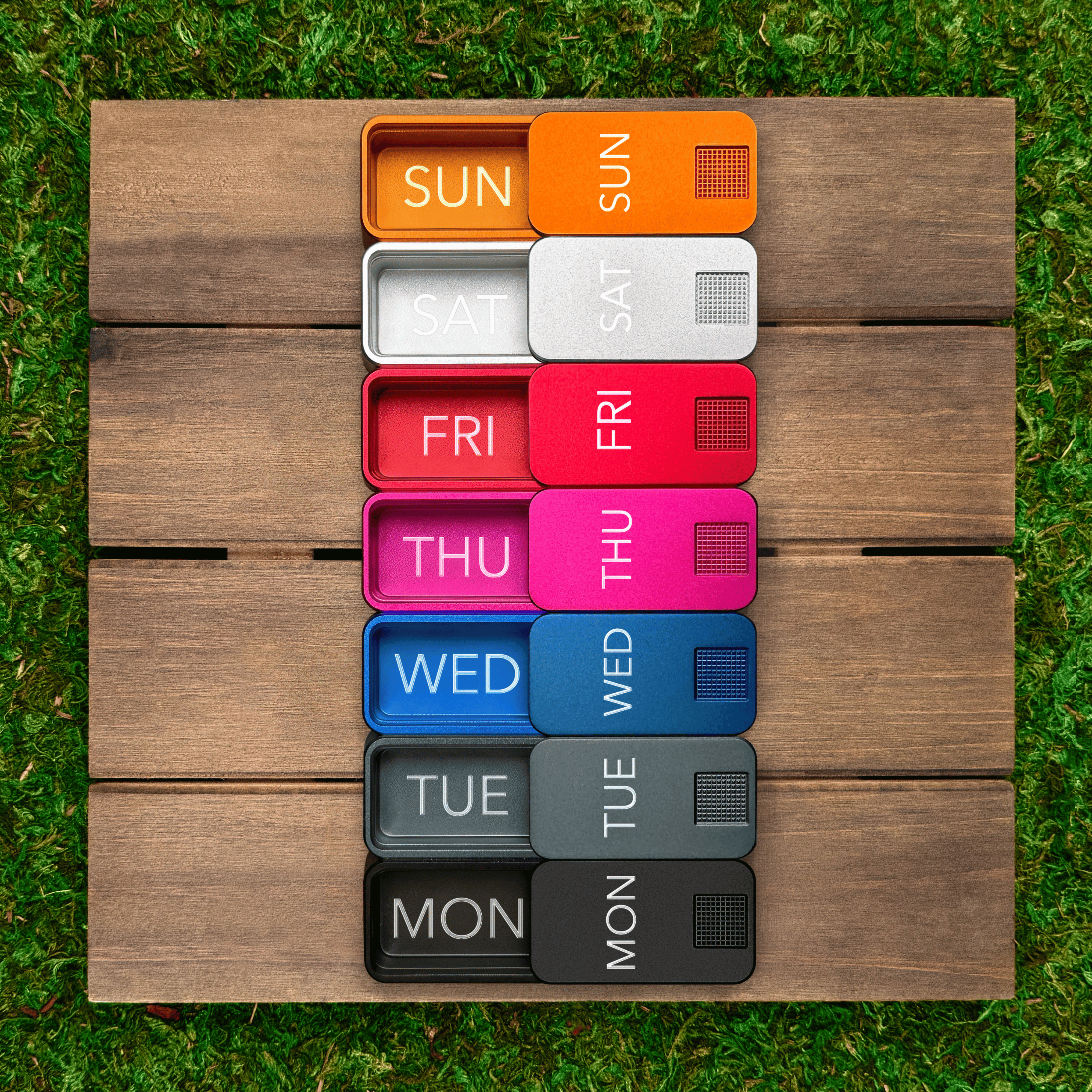 A colorful, weekly pill organizer with labeled daily compartments sits on wooden planks over green grass.