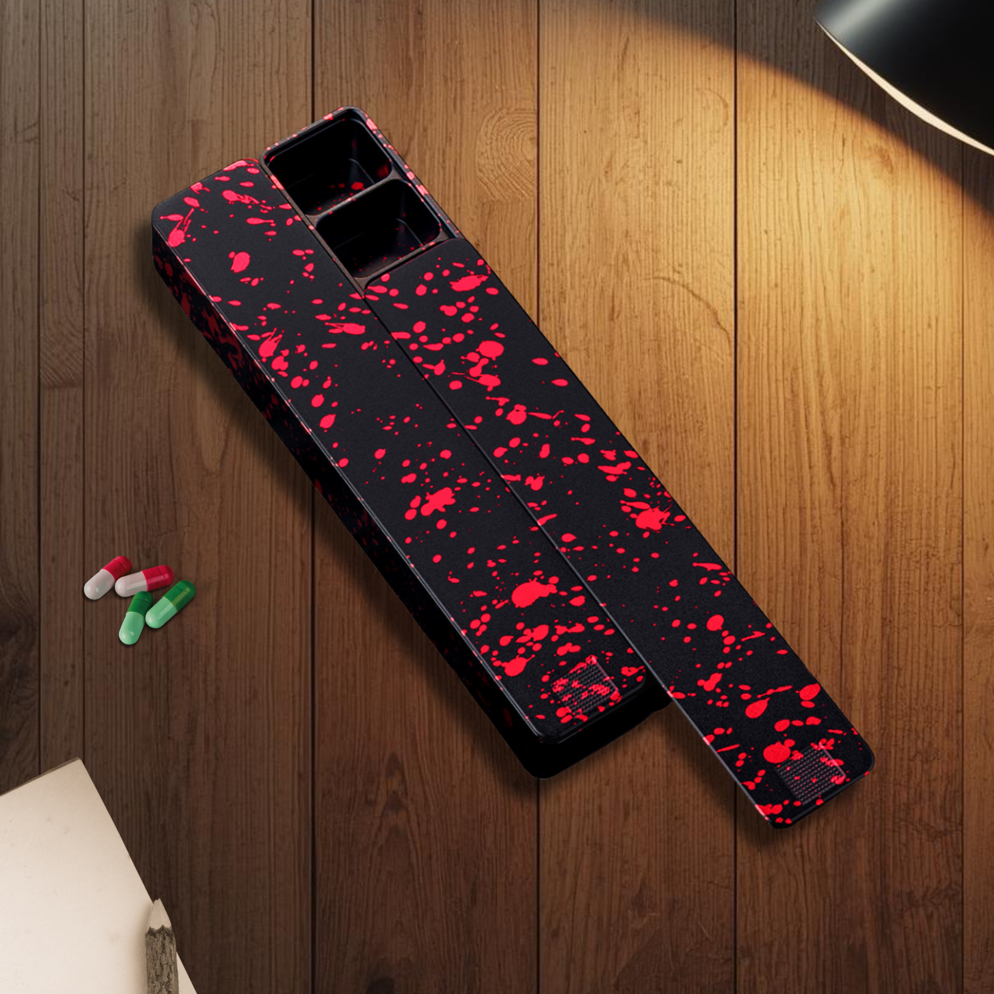 An open black pill organizer with a red splatter pattern sits on a wooden desk next to four pills.