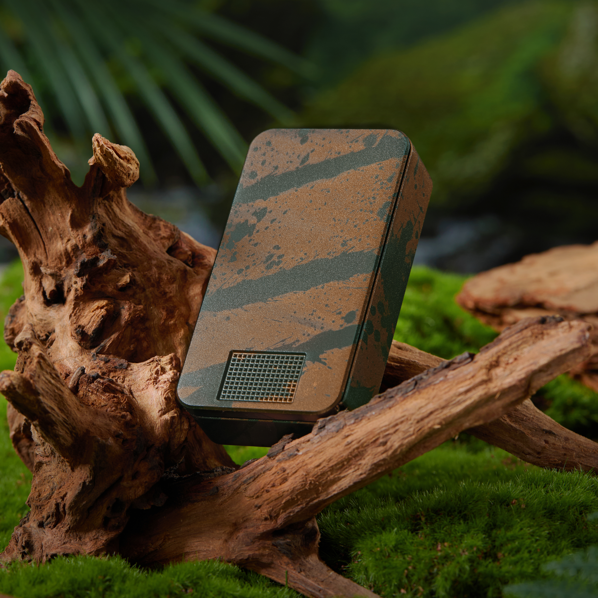 A rectangular device with a brown and green paint splatter pattern rests on driftwood and moss.