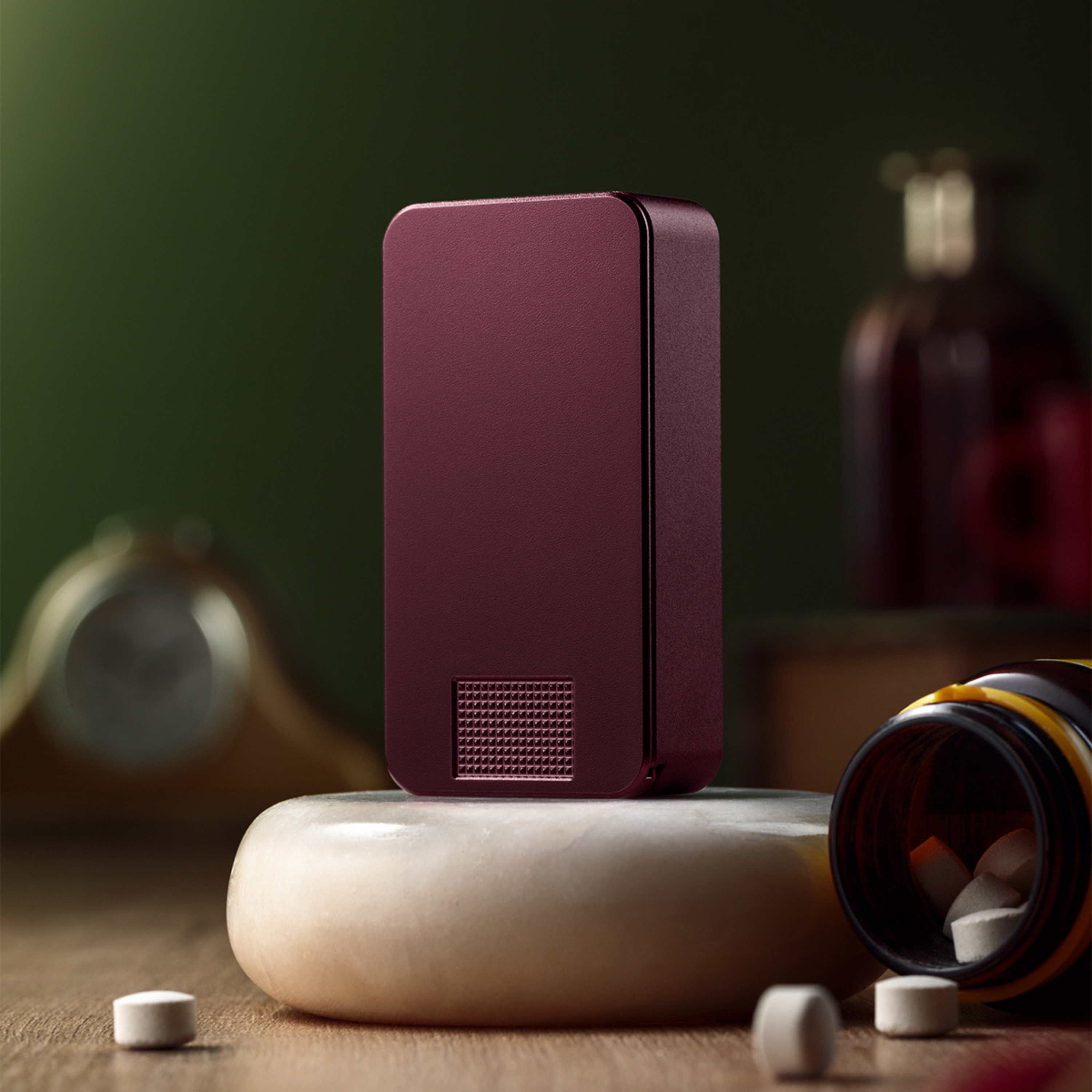 A maroon electronic device stands on a stone pedestal, surrounded by white pills and an open pill bottle.