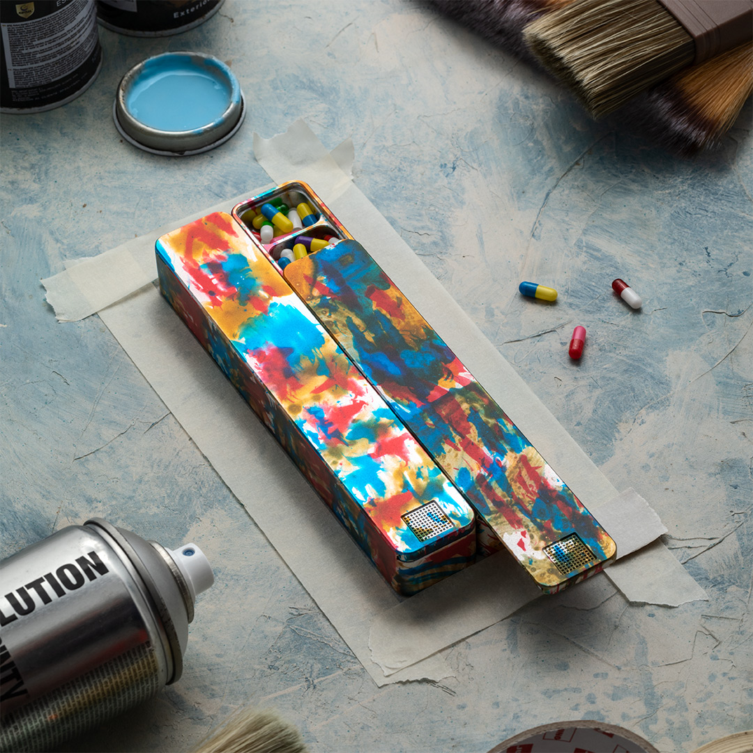 Colorful, painted box with pills, surrounded by paintbrushes and cans on a textured surface.