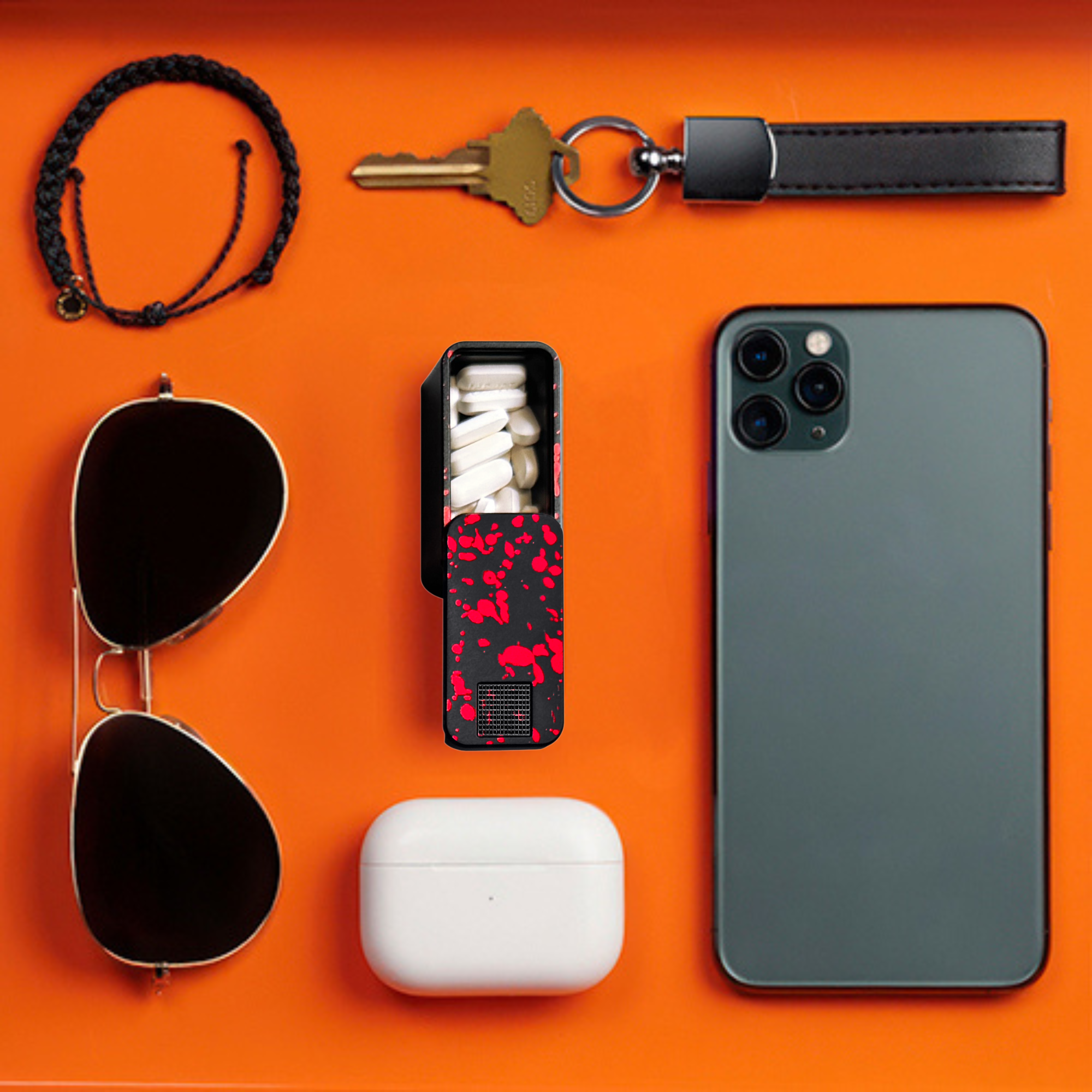 A flat lay of a phone, keys, sunglasses, earbuds, bracelet, and a pill case on an orange surface.