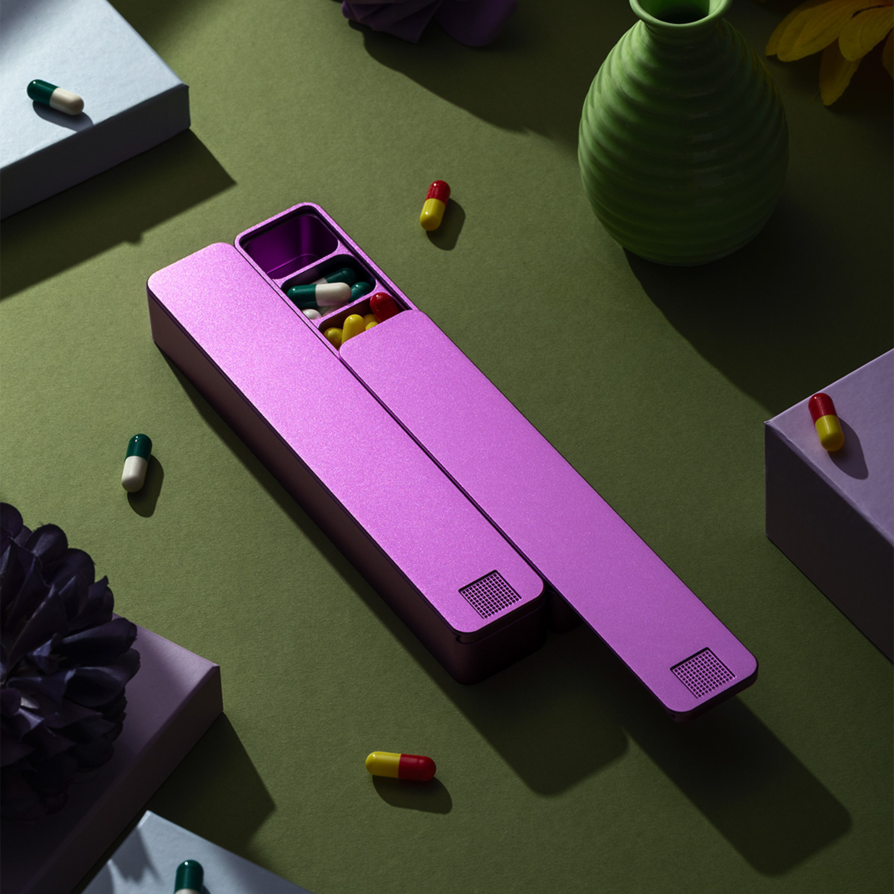 An open, metallic purple pillbox with colorful capsules is displayed on a green surface with decorative items.