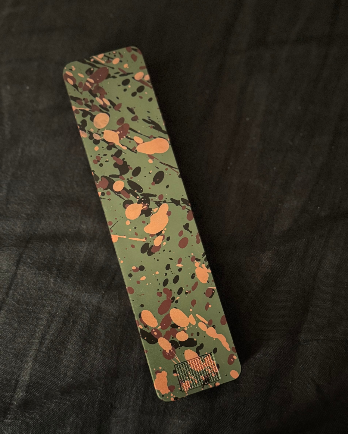 An olive green rectangular object with brown, black, and copper paint splatters on a black fabric background.