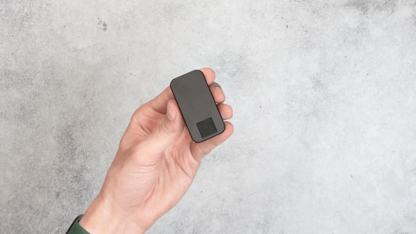 Hand holding a small black rectangular device against a gray background.
