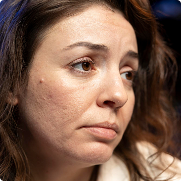 A close-up of a woman with brown hair and eyes looking away from the camera with a somber expression.