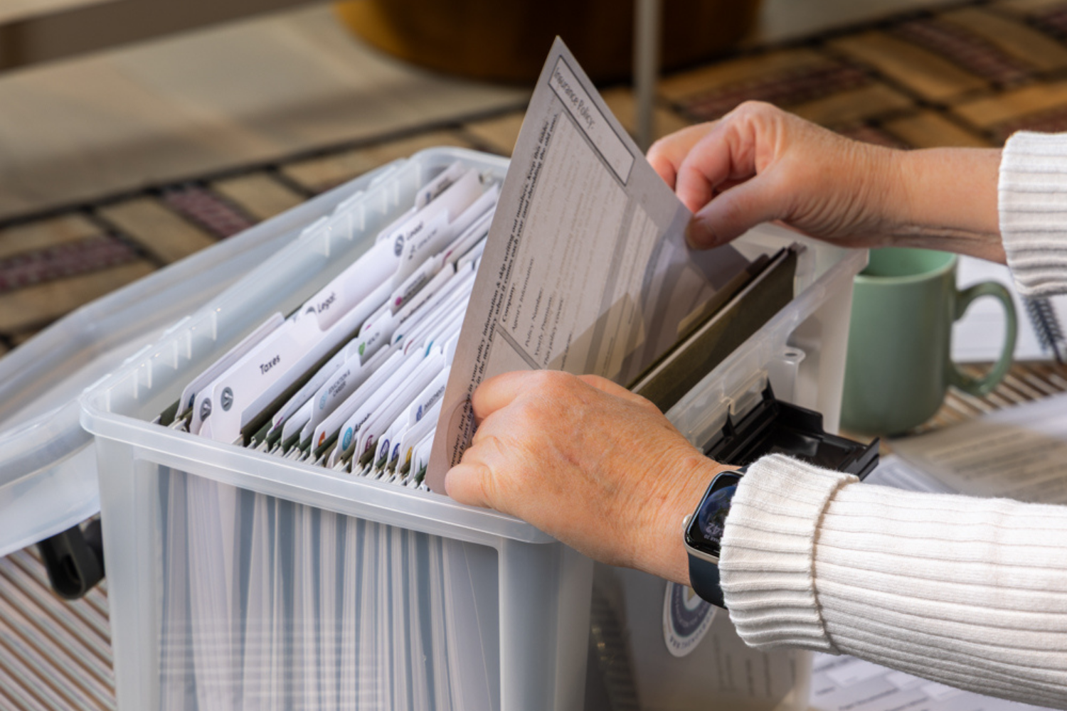 A person's hands file a document labeled 'Insurance Policy' into a clear plastic file box.