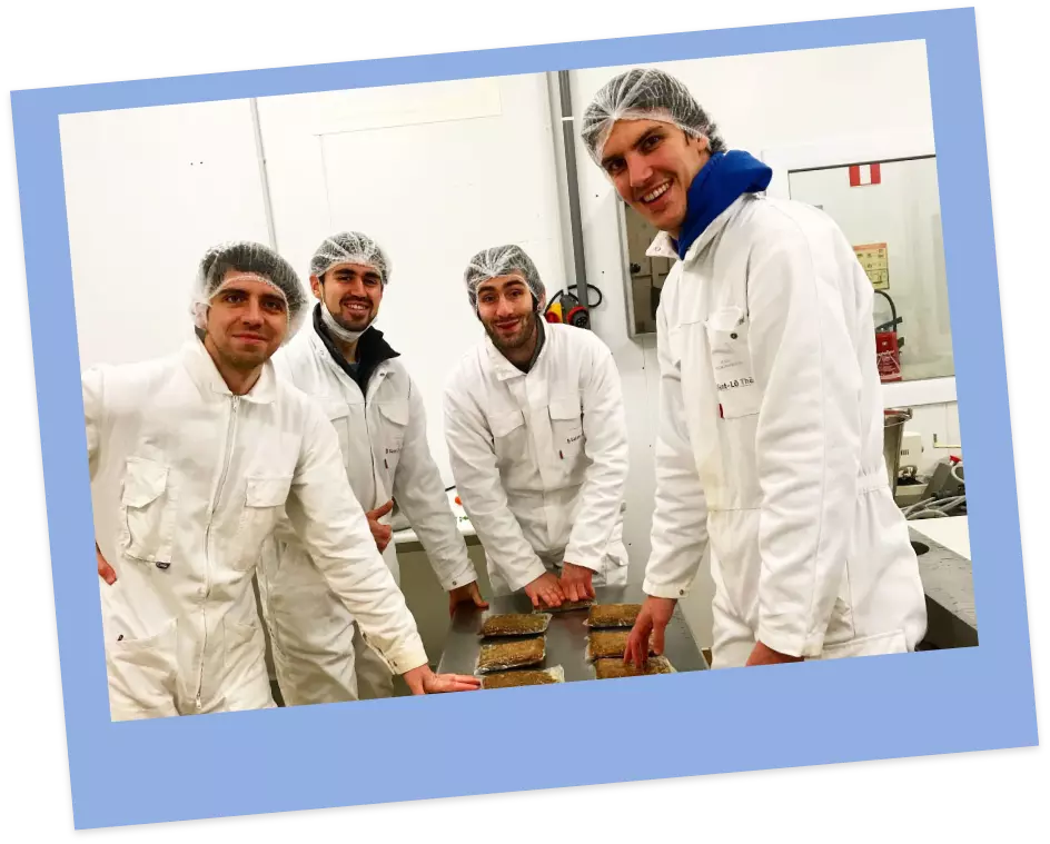 Four men in white coveralls and hairnets smile for a photo in a food production facility.