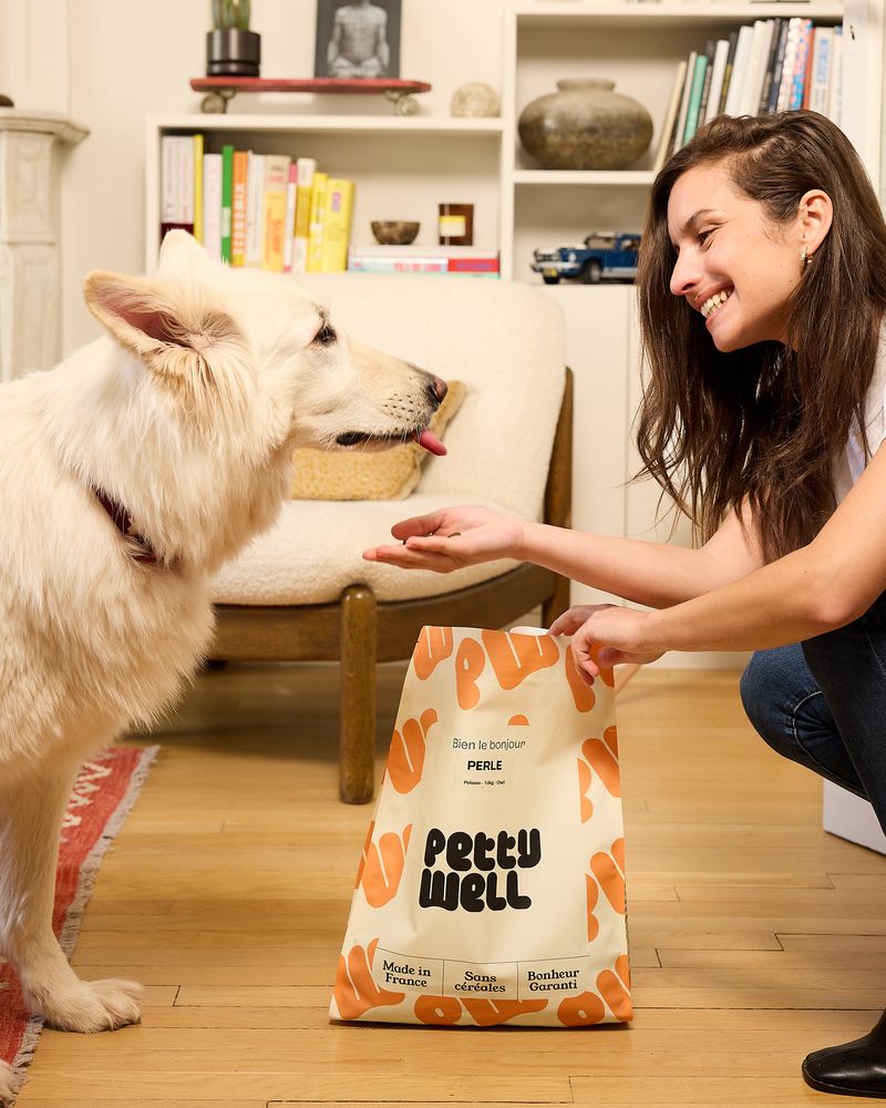 Woman smiling at a white dog with a pet food bag labeled 'Petty Well'.