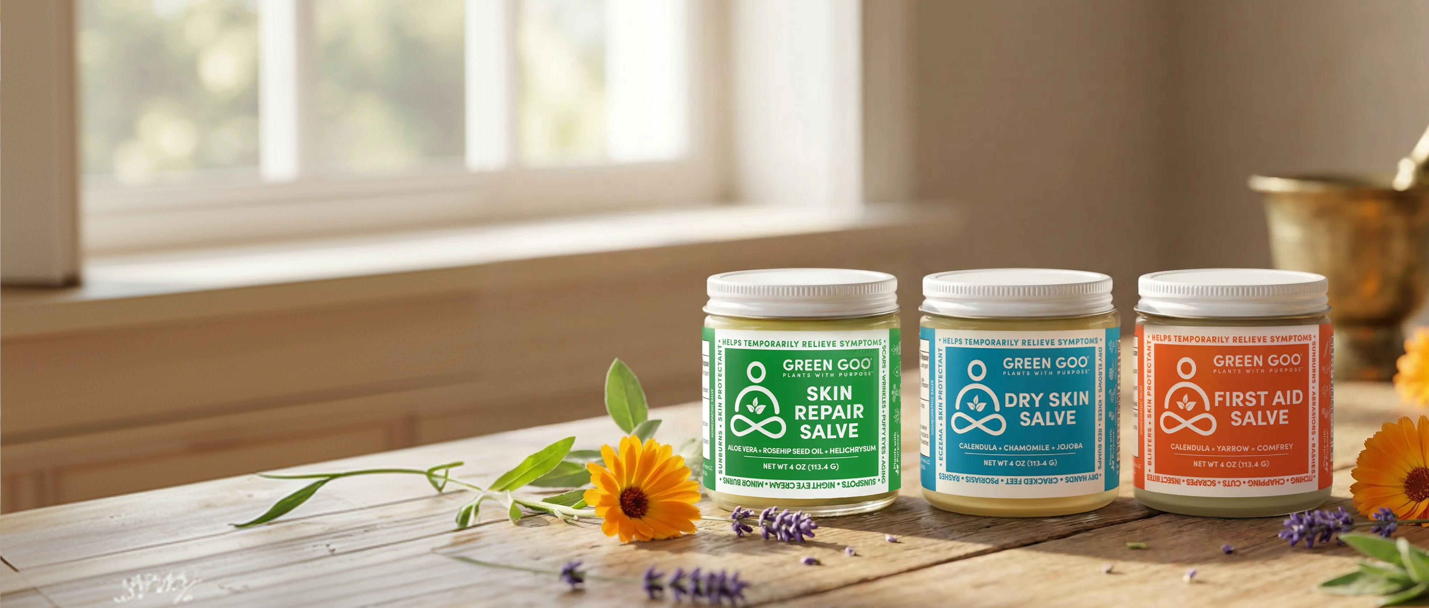 Three jars of Green Goo salves sit on a wooden table next to calendula and lavender flowers.