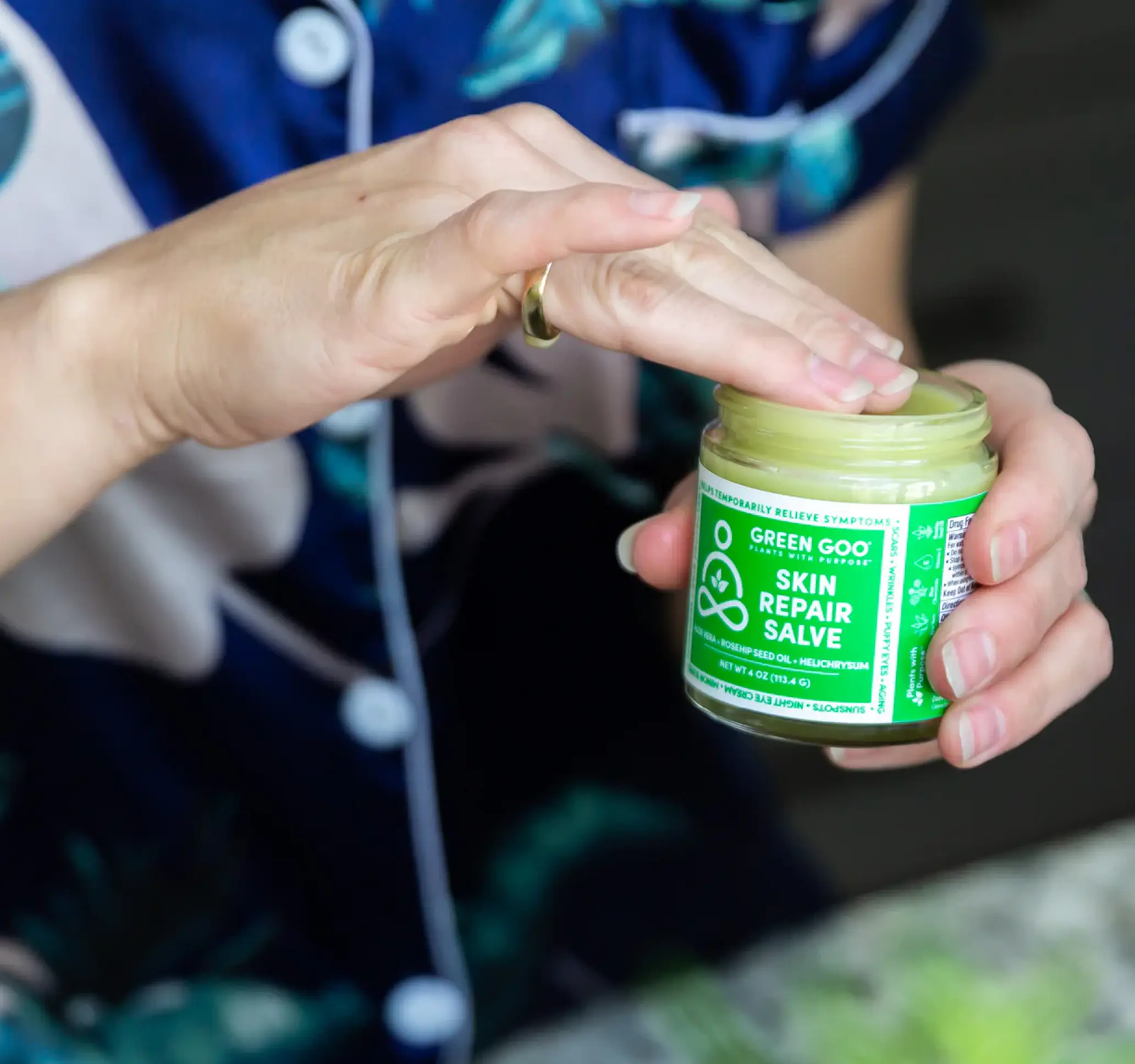 Hand dipping into a jar of skin repair salve labeled 'Green Goo'.