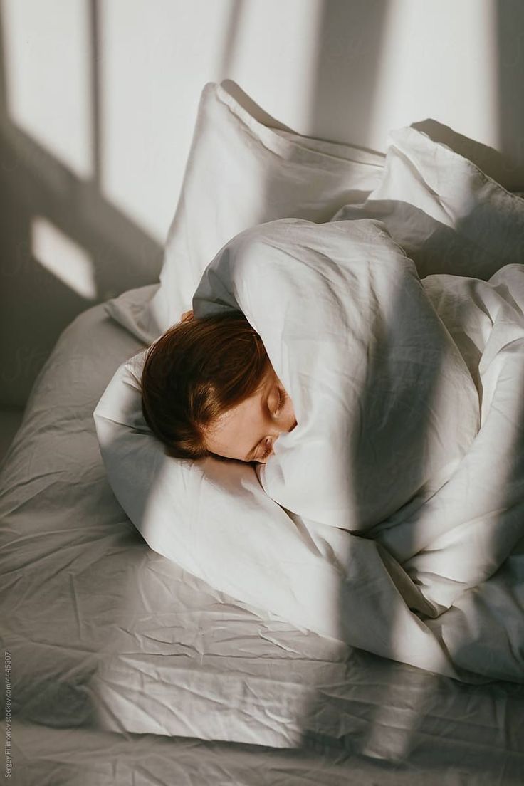 Person sleeping under white blankets with sunlight casting shadows.