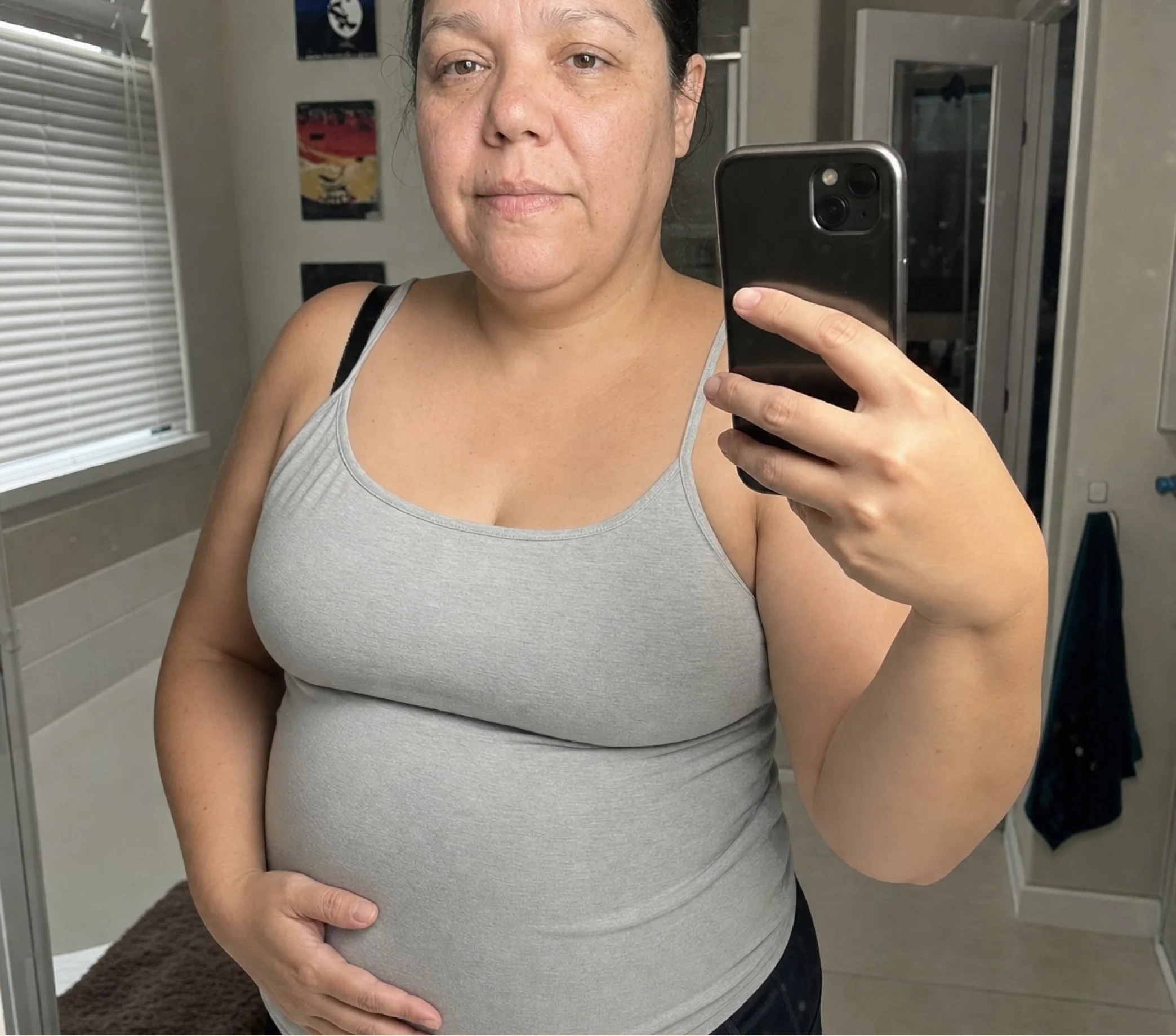 A woman in a grey tank top takes a mirror selfie with her smartphone, holding her stomach.