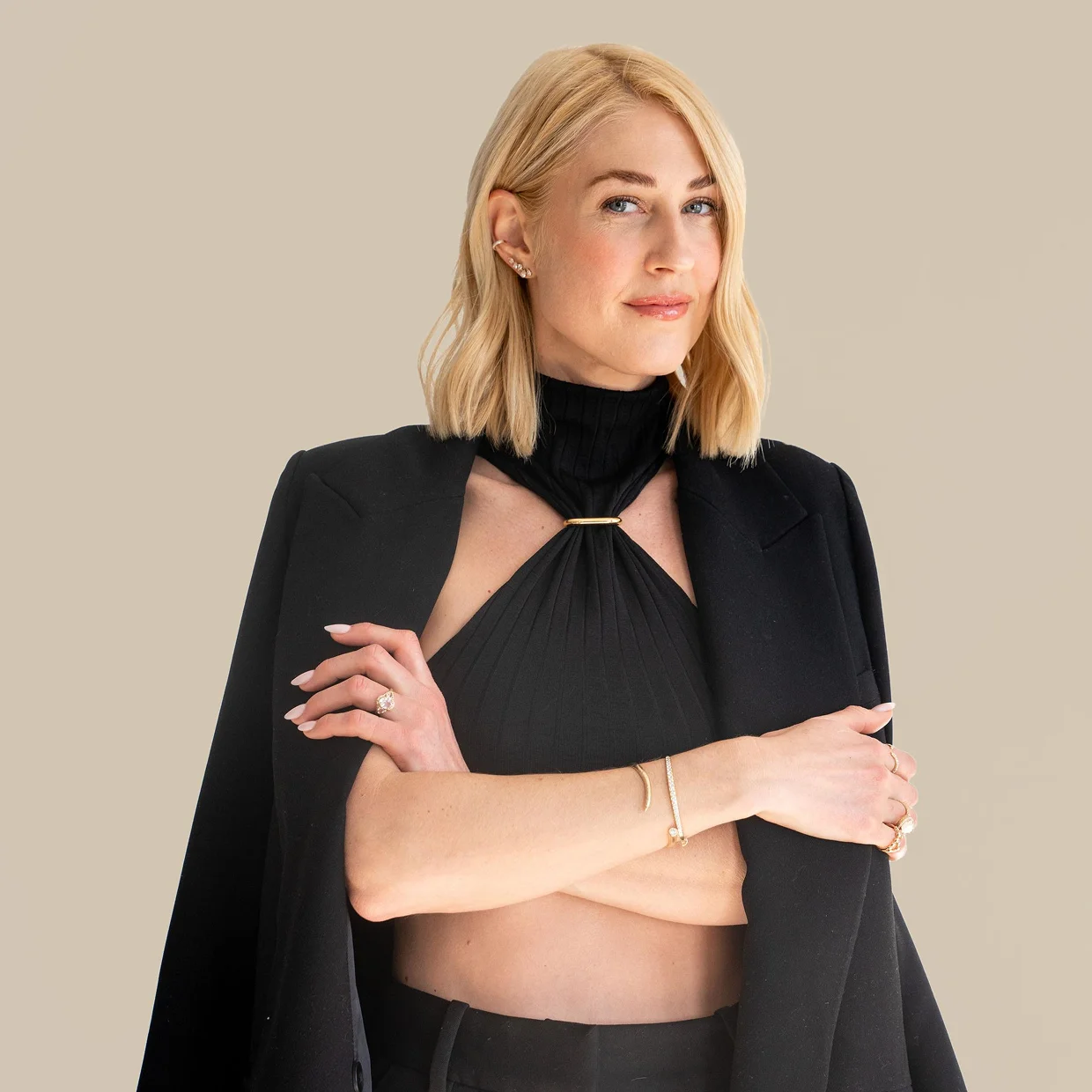 A woman with blonde hair in a black outfit with a blazer over her shoulders, arms crossed.