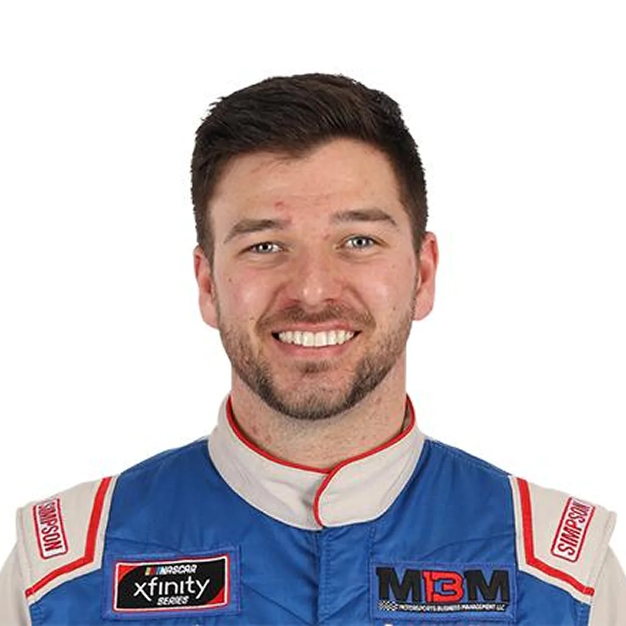 A smiling man with short brown hair and a beard wearing a blue, white, and red racing suit.