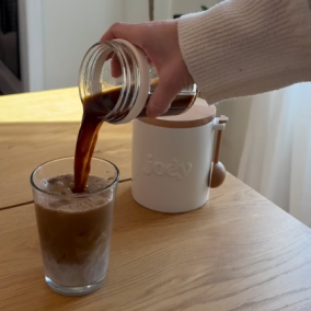 Hand pouring coffee into a glass with milk on a wooden table.