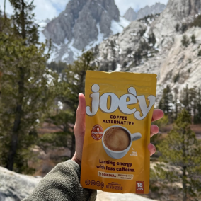 Hand holding a coffee alternative package in a mountainous landscape.