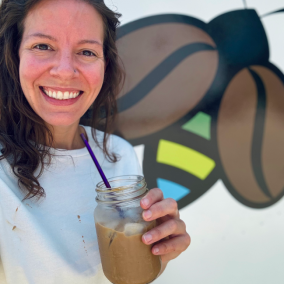 Person smiling, holding iced coffee in front of a bee mural.
