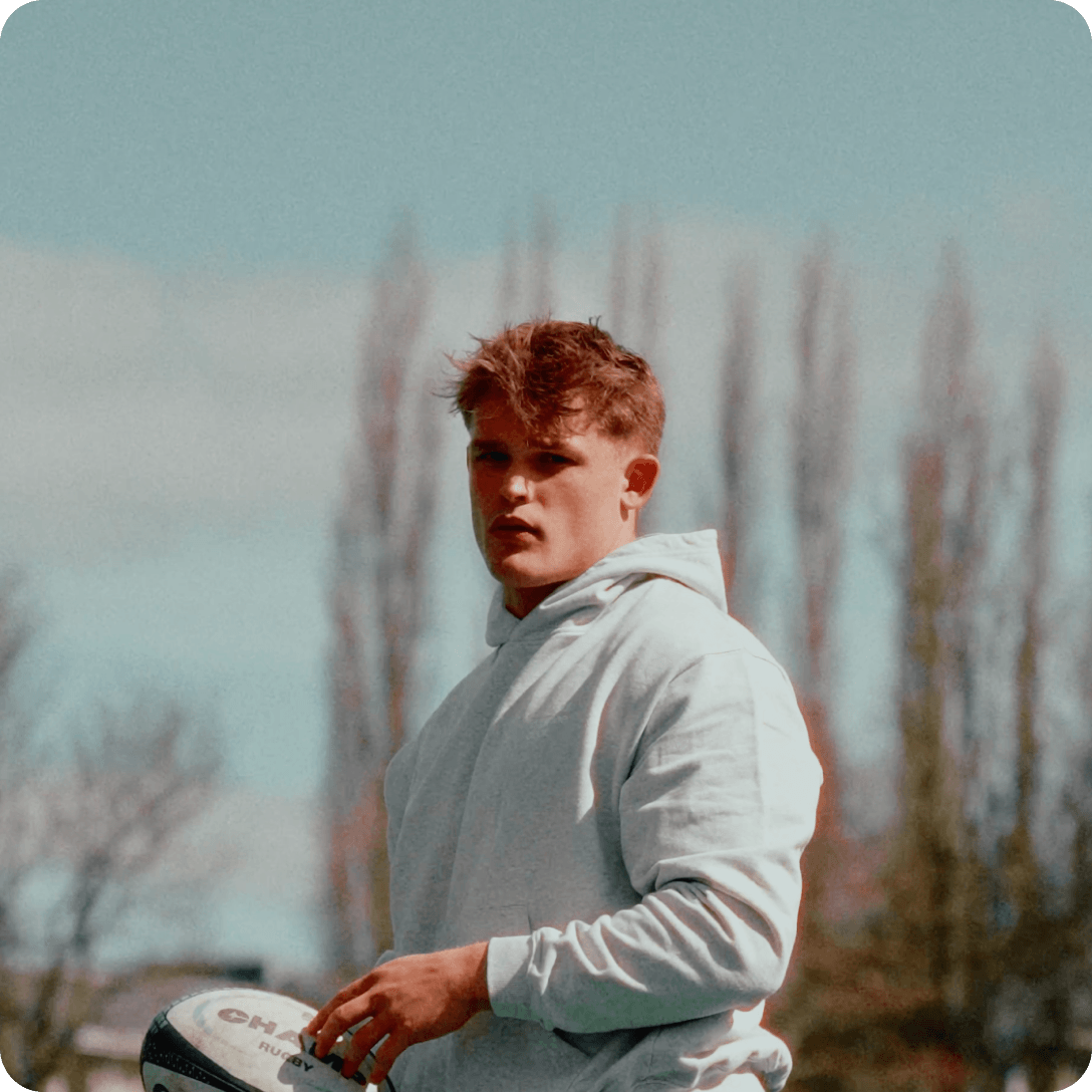 A young man in a light-colored hoodie stands outdoors holding a rugby ball and looking at the camera.