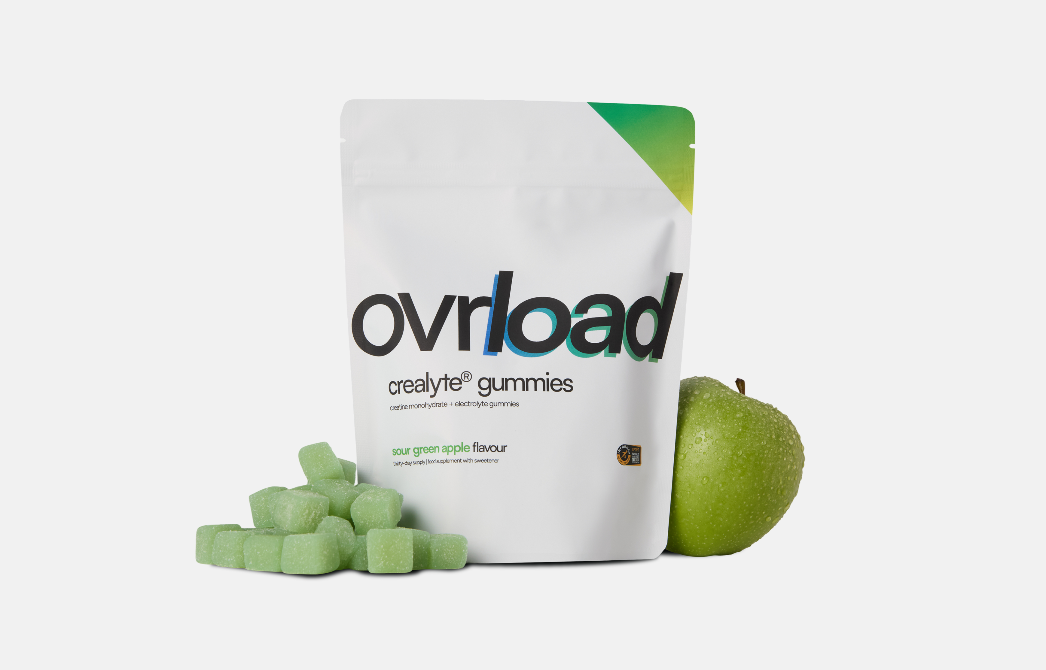 A white pouch of Ovrload sour green apple gummies next to a fresh green apple and loose gummies.