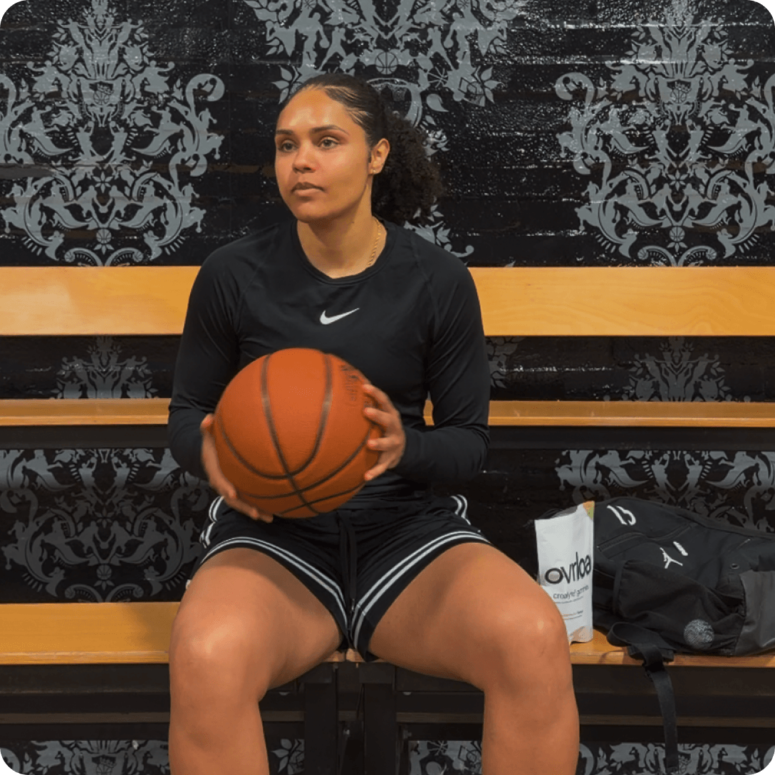A female basketball player in black athletic wear sits on a bench, holding a basketball.