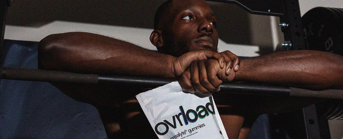 A shirtless man in a gym rests his arms on a bar, holding a pouch of Ovraload gummies.