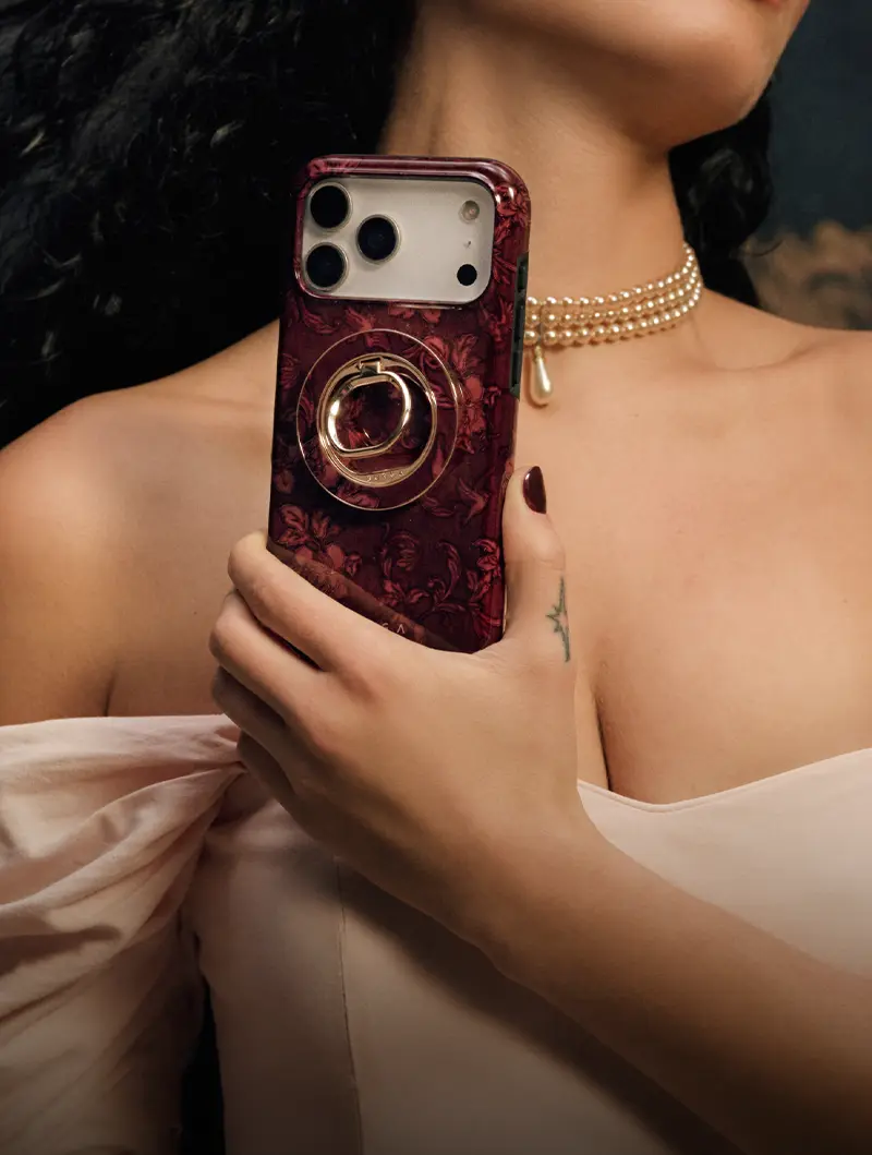 A woman wearing a pearl choker holds a smartphone in a red floral case with a gold ring.