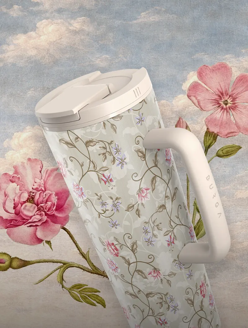 A travel mug with a vintage floral pattern and white handle against a painted floral and sky background.