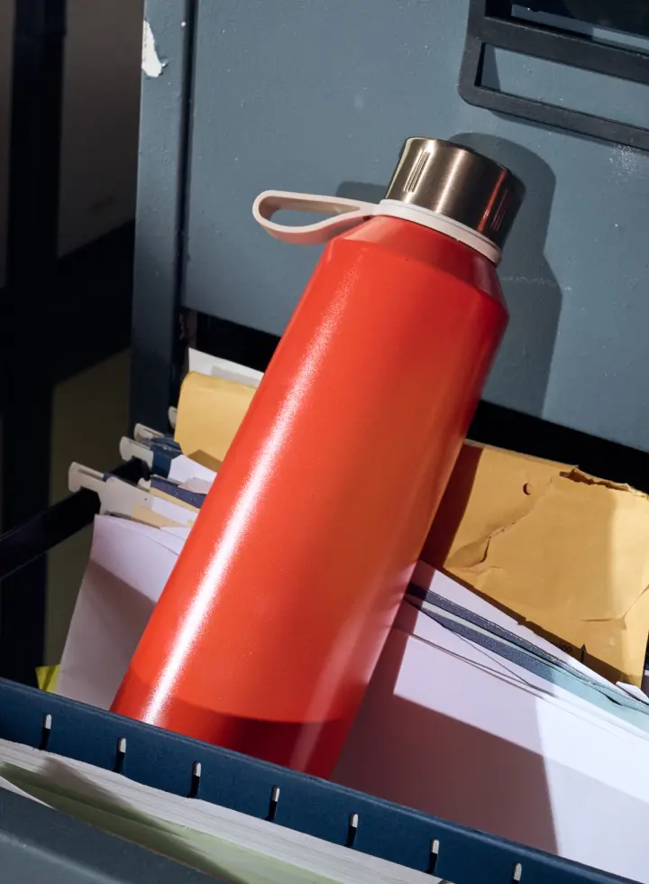 A red water bottle with a silver cap resting on top of files in an open filing cabinet drawer.