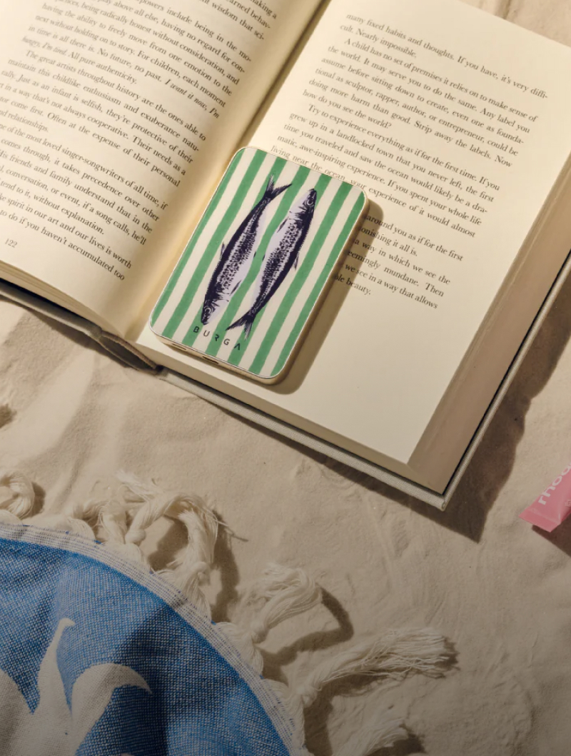 An open book with a striped, fish-design power bank on it, resting on sand next to a towel.