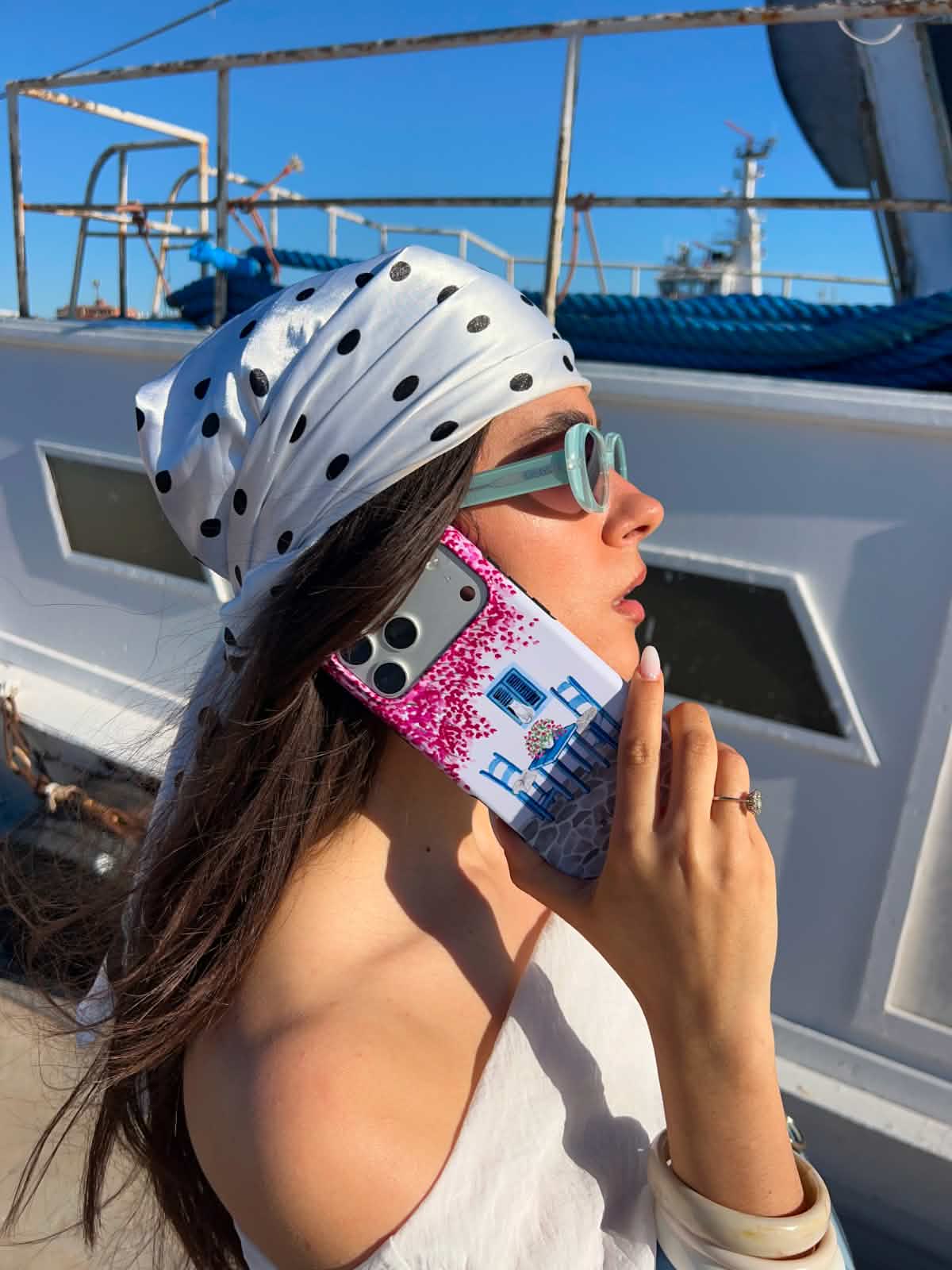 A woman in a polka-dot headscarf and sunglasses talks on her phone next to a boat.