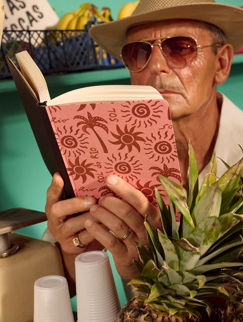 Man in sunglasses and hat reading a book near pineapples and plastic cups.