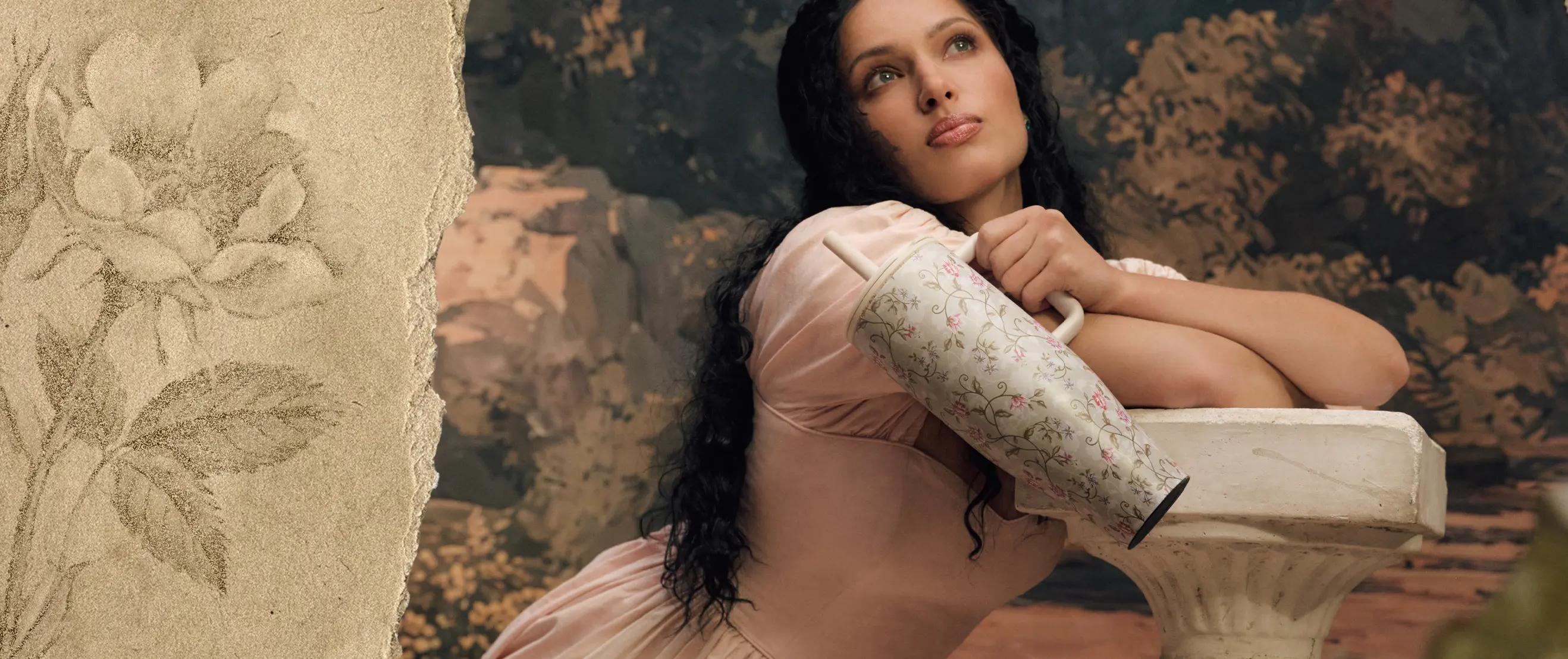 A woman in a pink dress leans on a pedestal, holding a floral tumbler and looking upwards.