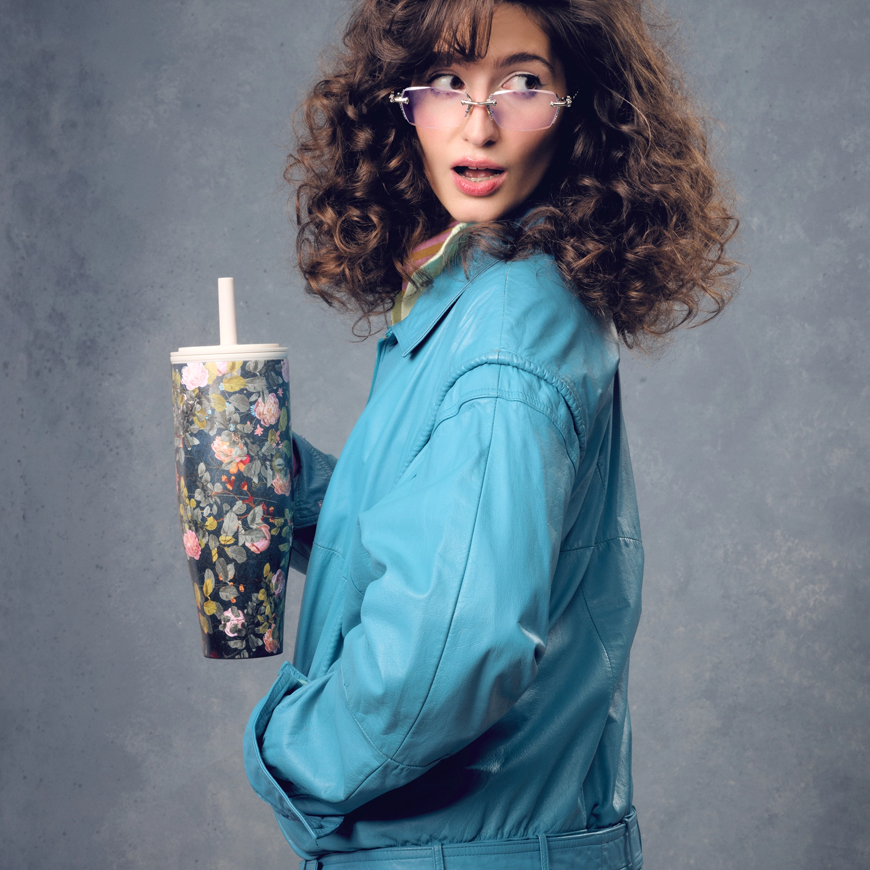 A woman in a blue jacket and glasses holds a floral cup while looking over her shoulder.