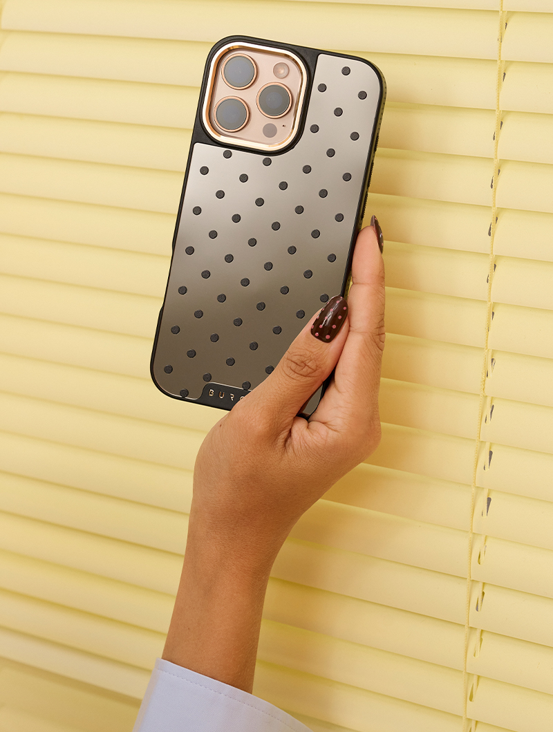Hand holding a polka-dotted phone case against light yellow blinds.