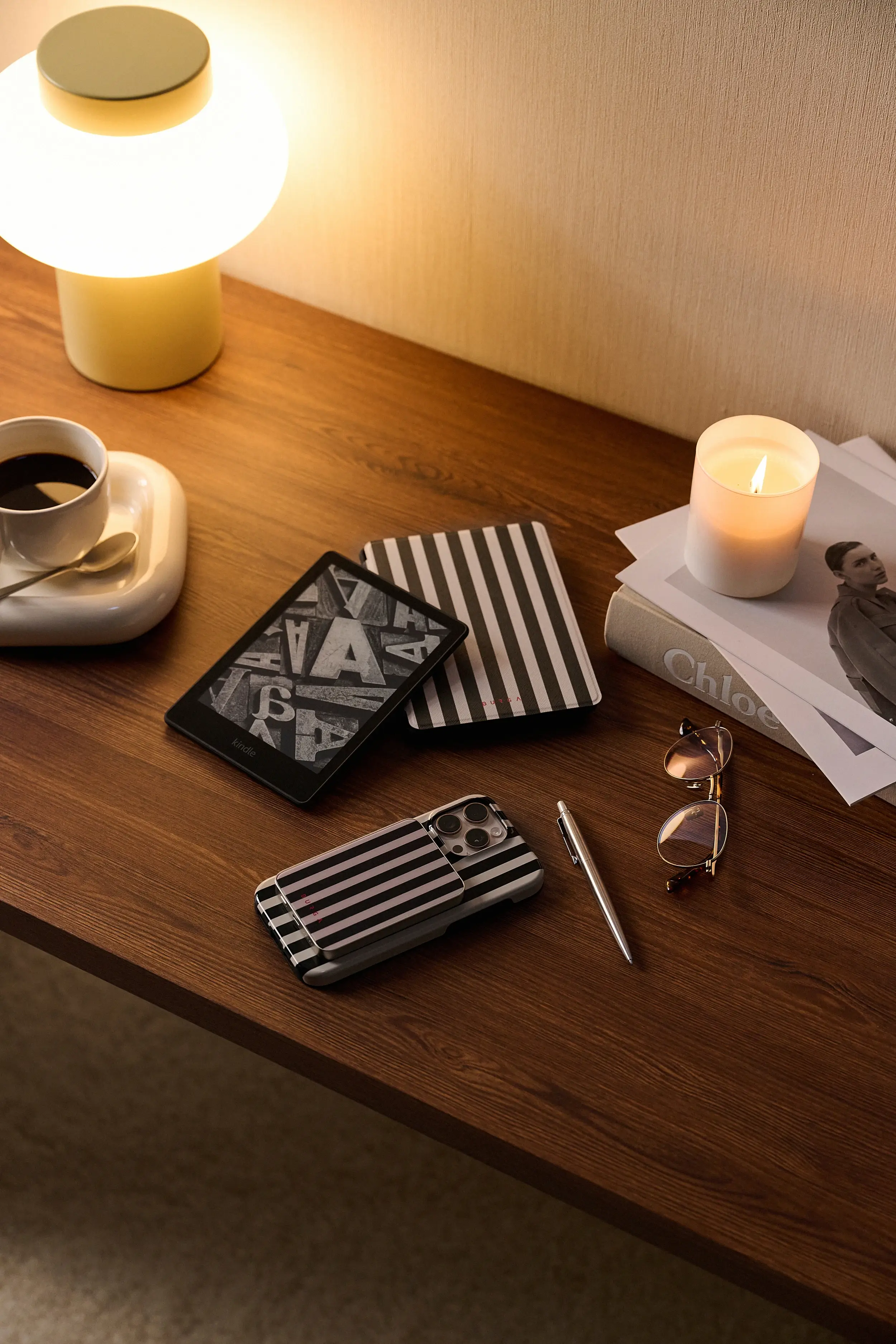 Wooden desk with a lamp, tablet, phone, candle, book, glasses, and coffee cup.