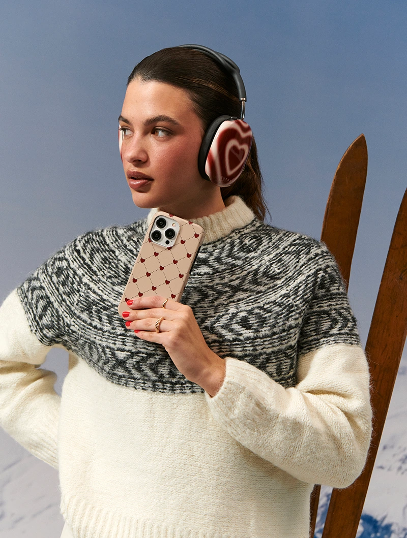 Model in a Nordic sweater with skis, holding a heart-pattern phone case and wearing heart headphone covers