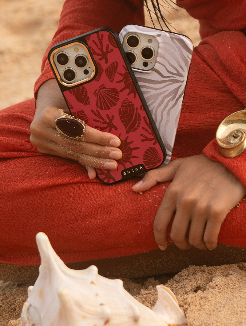 Person holding two patterned phone cases on a sandy beach.