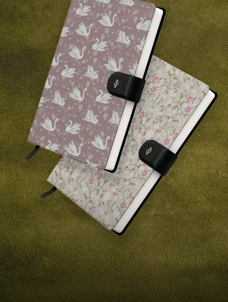 An overhead view of two patterned notebooks, one with swans and one with flowers, on a green surface.