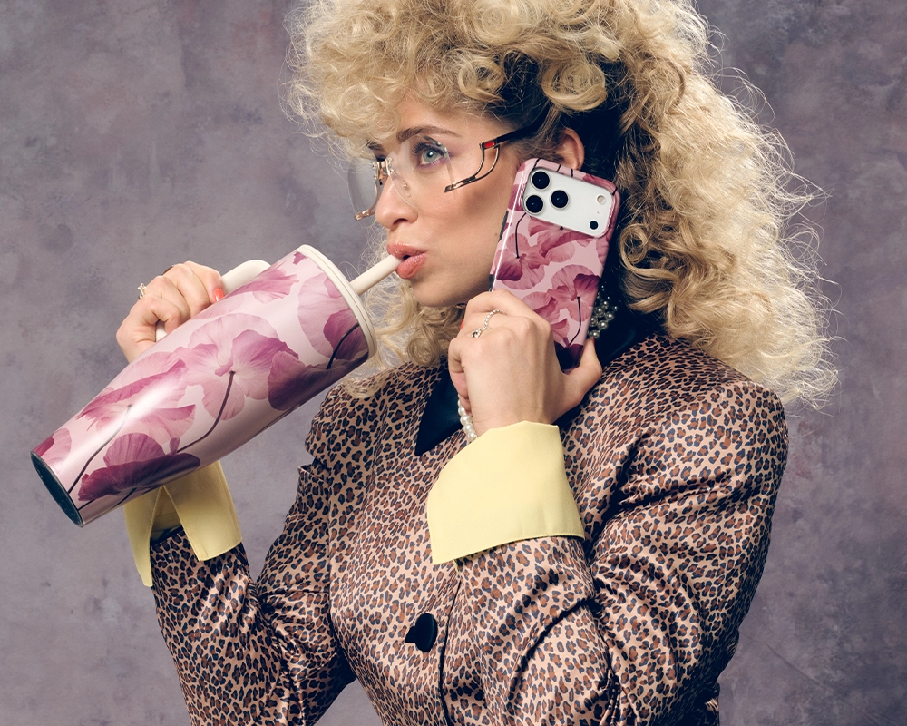 A woman with curly hair holds a floral phone and drinks from a matching tumbler.