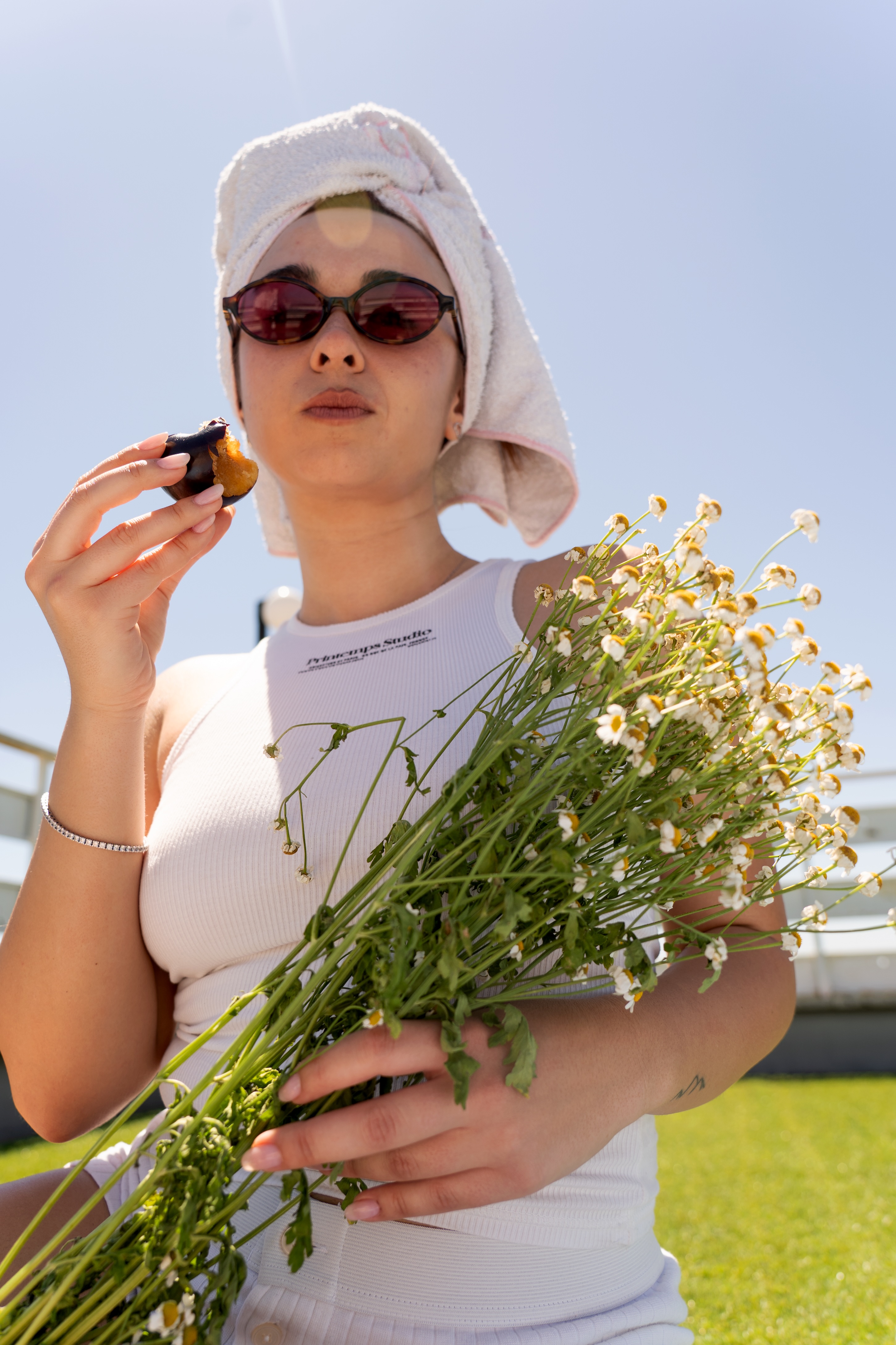 Ritratto di una persona con un asciugamano sui capelli e occhiali da sole, che tiene in mano un piccolo contenitore di prodotto e un grande mazzo di camomilla, simboleggiando un rituale haircare naturale e delicato di Originae, con trattamenti pensati per il cuoio capelluto e la pelle.