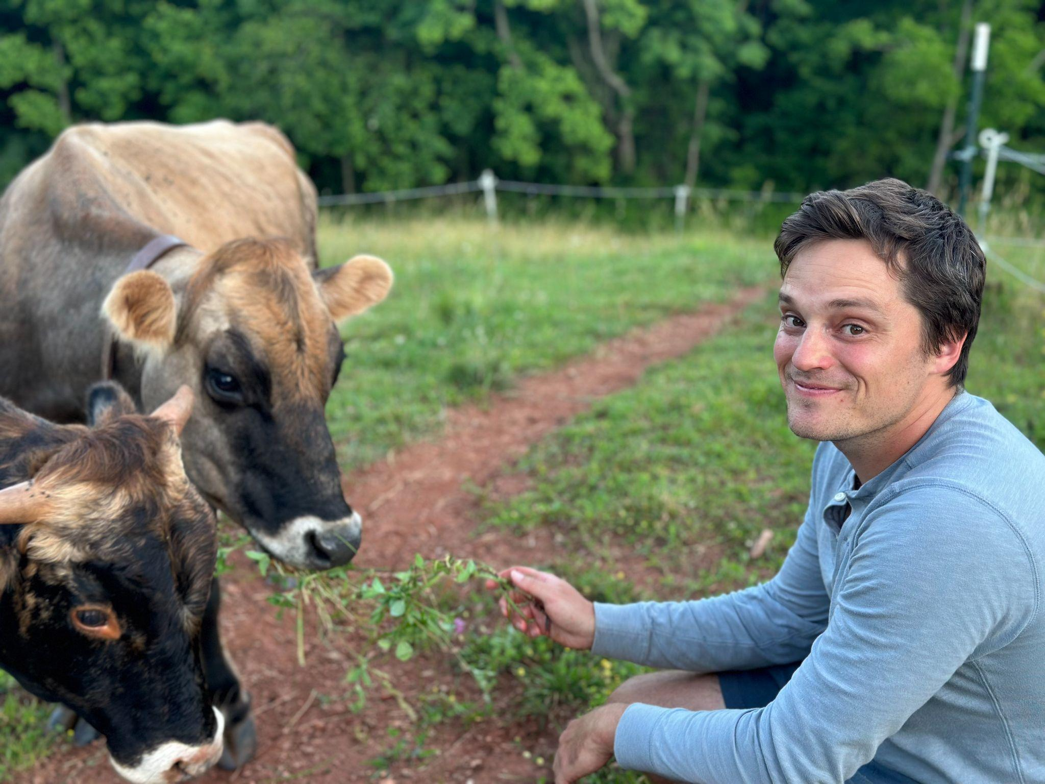 Person smiling, crouching beside two cows, in a grassy field.