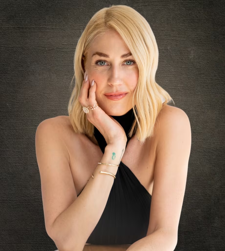 Person with blonde hair in a black outfit, smiling with hand on chin.